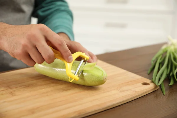 Man peeling zucchini at kitchen table, closeup. Preparing vegetable