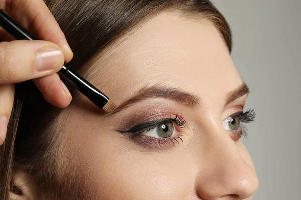 Artist correcting woman\'s eyebrow shape with powder on grey background, closeup