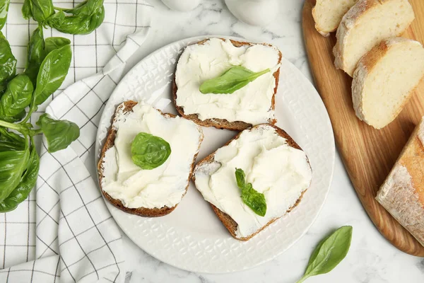 Bread with cream cheese and basil on white marble table, flat lay