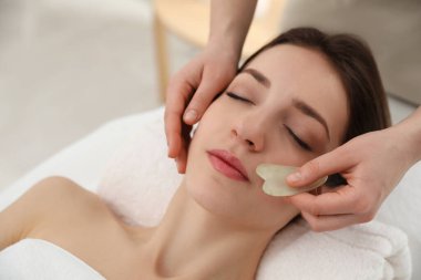 Genç bir kadın güzellik salonunda gua sha aracıyla yüz masajı yaptırıyor.