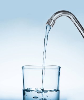 Açık mavi arkaplandaki musluktan gelen suyla doldurma bardağı
