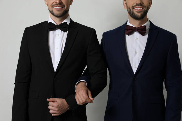 Happy gay couple in elegant suits standing near white wall, closeup
