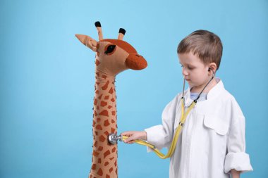 Pediatri üniformalı şirin bir çocuk steteskopla ve açık mavi arka planda oyuncak zürafayla oynuyor.