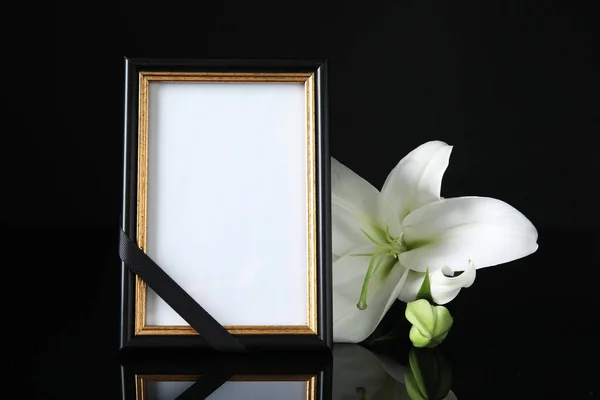 Funeral photo frame with ribbon and white lily on black table against dark background. Space for design
