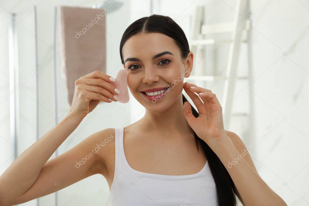 Beautiful young woman doing facial massage with gua sha tool in bathroom