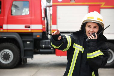 İtfaiyeci üniformalı. Dışarıdaki itfaiye aracının yanında taşınabilir telsiz kullanıyor.