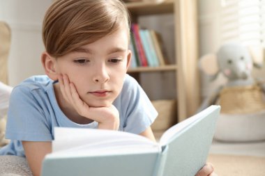 Küçük çocuk evde yerde kitap okuyor.