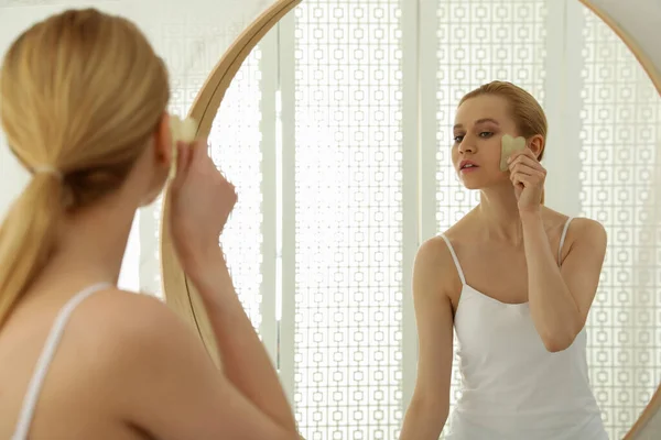 漂亮的年轻女子在家里用瓜沙工具在镜子前做面部按摩 — 图库照片