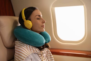 Seyahat yastığı olan genç bir kadın uçuş sırasında uçakta müzik dinlerken dinleniyor.