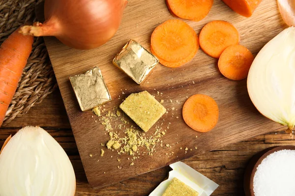 Bouillon cubes, cut carrot and onions on wooden table, flat lay