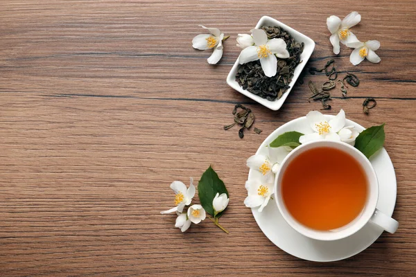 Cup of aromatic jasmine tea, dry leaves and fresh flowers on wooden table, flat lay. Space for text