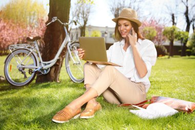 Parkta dizüstü bilgisayarla çalışan bir kadın akıllı telefondan konuşuyor.