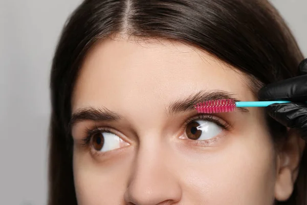 Beautician brushing woman's eyebrows before tinting on grey background, closeup