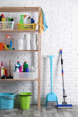 Shelving unit with detergents, cleaning tools and toilet paper near white brick wall indoors