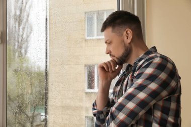 Depressed man near window at home. Space for text clipart