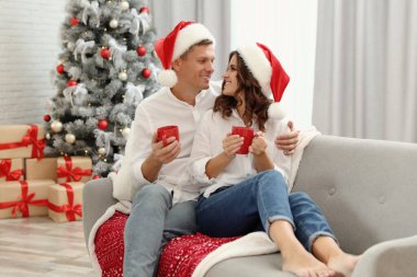 Noel Baba şapkalı mutlu çift evde Noel ağacının yanında bir bardak sıcak içecekle