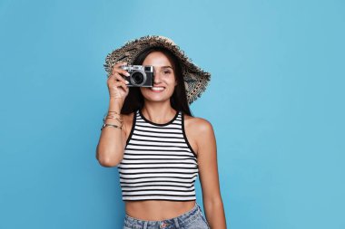 Beautiful young woman with straw hat and camera on light blue background