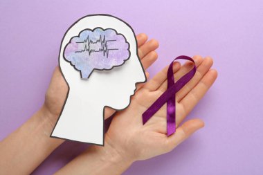 Woman holding human head cutout, brain and purple ribbon at violet background, top view. Epilepsy awareness