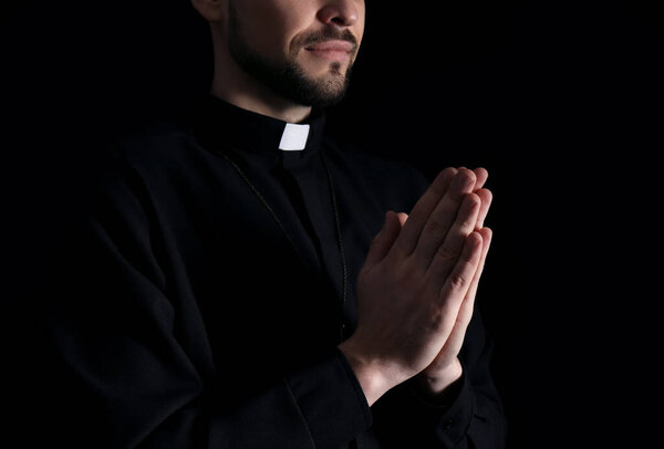 Priest in cassock praying on dark background, closeup