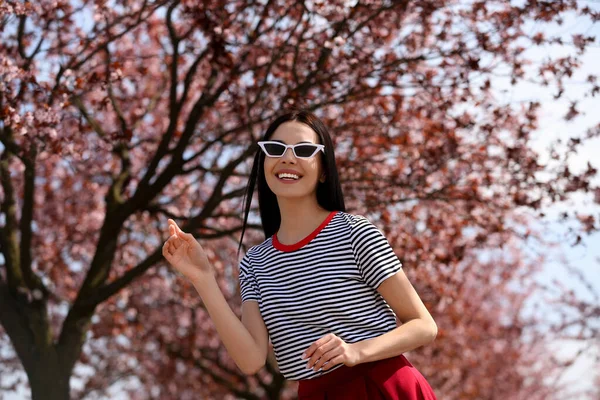 屋外の美しい開花木の近くにサングラスを持つかなり若い女性 スタイリッシュな春の表情 — ストック写真