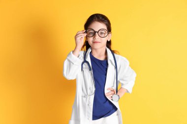 Sarı arka planda doktor gibi giyinmiş gözlüklü ve steteskoplu küçük kız. Çocuk doktoru pratiği.