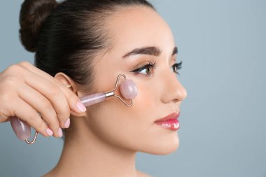 Gri arka planda doğal pembe kuvars yüz silindiri kullanan kadın, yakın plan. Metin için boşluk