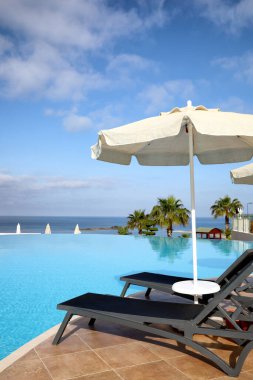Tatil köyündeki sonsuz havuza yakın şezlonglar ve plaj şemsiyesi.