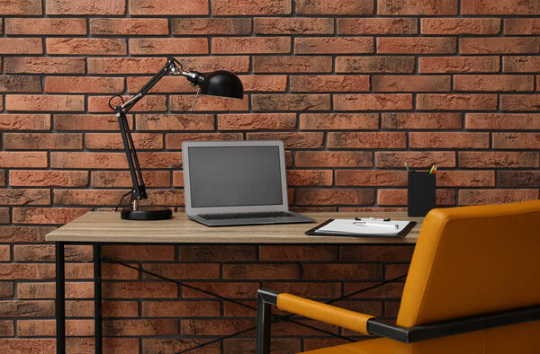 Stylish workplace with laptop and comfortable chair near brick wall. Interior design