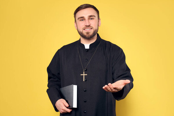 Priest in cassock with Bible reaching out his hand on yellow background