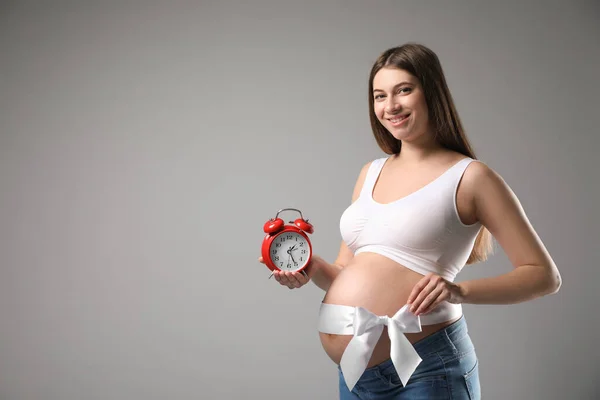 目覚まし時計と灰色の背景に弓を持つ若い妊婦 テキストのためのスペース 出産までの時間 — ストック写真