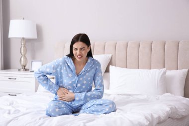 Young woman suffering from stomach ache in bedroom clipart