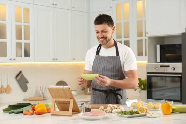 Happy man making dinner while watching online cooking course via tablet in kitchen