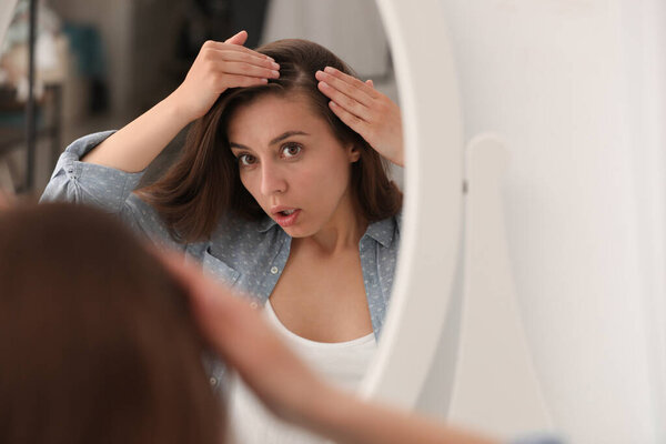 Emotional woman with hair loss problem looking in mirror indoors
