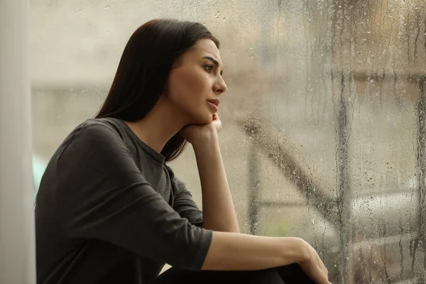 Depressed woman near window on rainy day, space for text