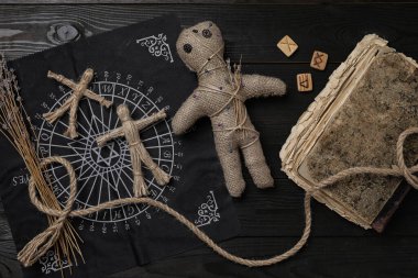 Voodoo doll with pins surrounded by ceremonial items on black wooden table, flat lay clipart