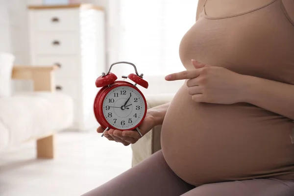 腹の近くの目覚まし時計を指して妊娠中の若い女性は屋内で閉じます 出産までの時間 — ストック写真