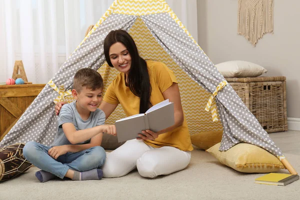 Mother and son reading book near toy wigwam at home
