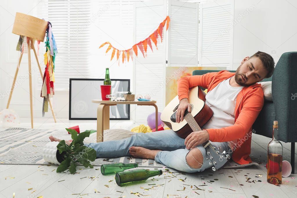 Young man with guitar sleeping near sofa in messy room after party