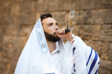 Jewish man blowing shofar on Rosh Hashanah outdoors. Wearing tallit with words Blessed Are You, Lord Our God, King Of The Universe, Who Has Sanctified Us With His Commandments, And Commanded Us To Enwrap Ourselves In Tzitziton in Hebrew clipart