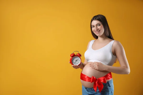 目覚まし時計と黄色の背景に赤い弓を持つ若い妊婦 テキストのためのスペース 出産までの時間 — ストック写真