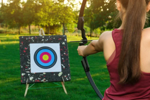 Woman with bow and arrow aiming at archery target in park, back view