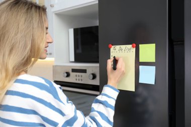 Genç bir kadın mutfaktaki buzdolabının kapısına liste yapmak için yazıyor.