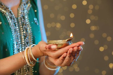 Kahverengi arka planda yanan diya lambasını tutan, yakın görüşlü, bulanık ışıklı bir kadın. Diwali tatili