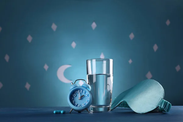Alarm clock, soporific pills and sleeping mask near glass of water on table against blue wall decorated with stars and crescent, space for text. Insomnia treatment