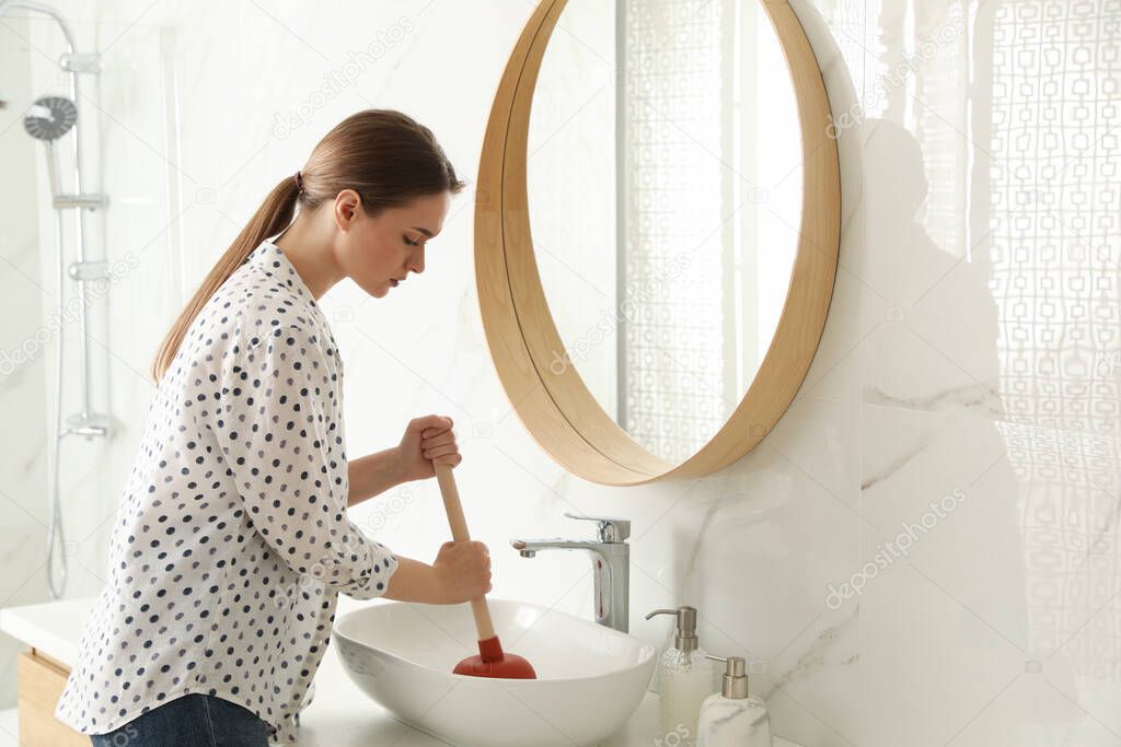 Young woman using plunger to unclog sink drain in bathroom
