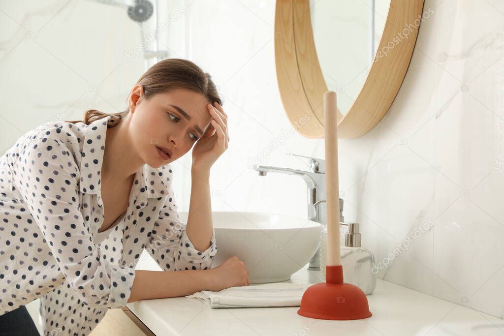 Unhappy young woman with plunger near clogged sink in bathroom