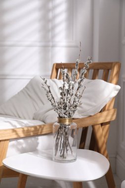 Glass vase with pussy willow tree branches on table near armchair in room