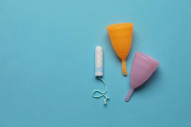 Açık mavi arka planda tampon yakınında adet bardakları, metin için yeri olan düz yataklar. Yeniden kullanılabilir ve tek kullanımlık kadın ürünleri
