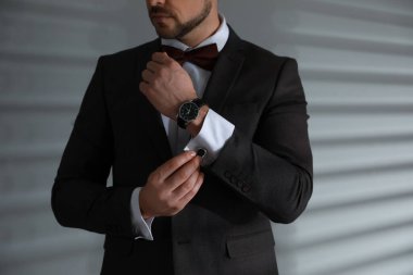 Beyaz duvarın yanında şık takım elbiseli ve kol düğmeli bir adam var.