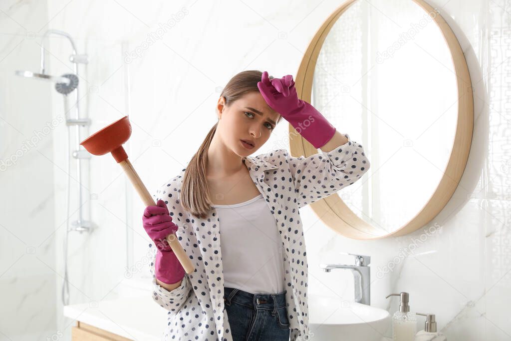 Tired young woman with plunger near sink in bathroom
